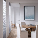 A framed square art print depicting a calm blue sea with sailboats, hung on a white wall in a modern dining room with a wooden table and white chairs.